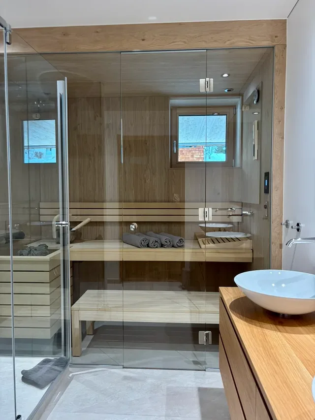 Moderne Sauna hinter Glaswänden mit Holzbänken, Towels und Fenster; daneben Waschbecken auf Holzkonz