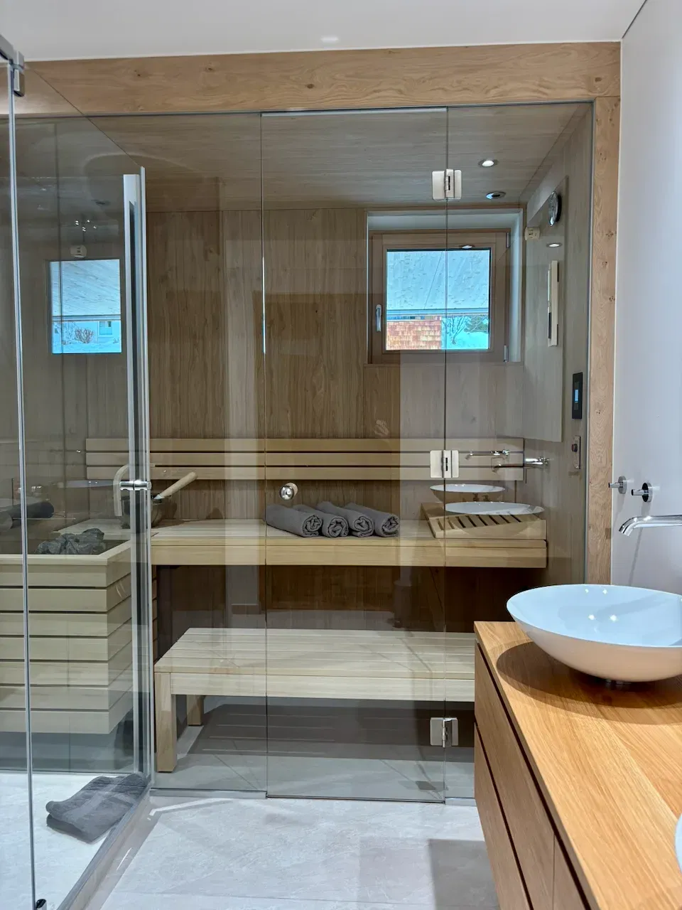 Moderne Sauna hinter Glaswänden mit Holzbänken, Towels und Fenster; daneben Waschbecken auf Holzkonz