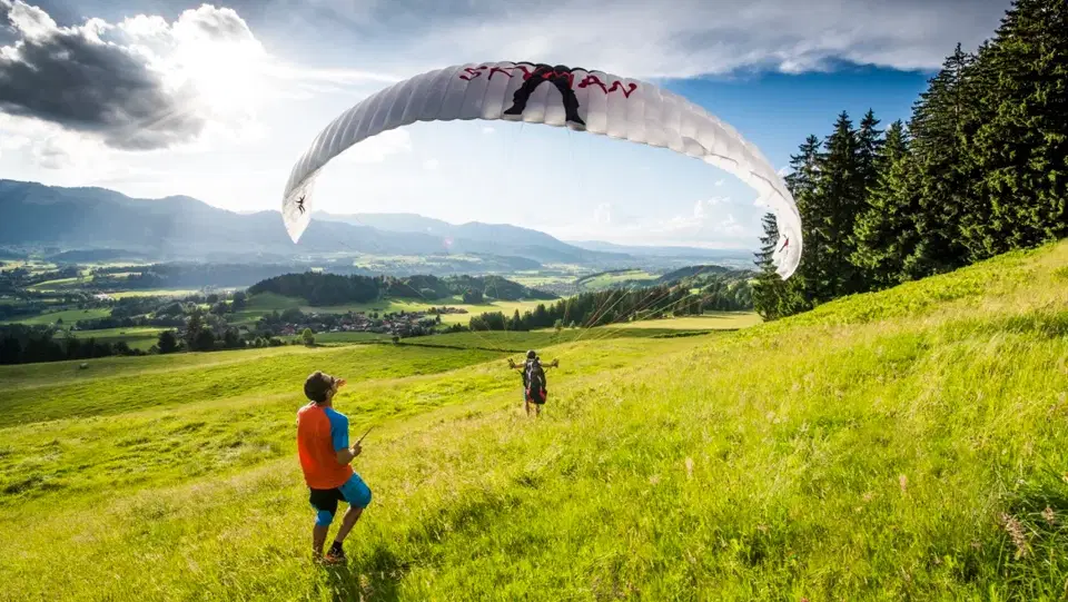 Paragliding-Szene auf grünem Hang; zwei Personen beobachten, Paraglider in der Luft über der Wiese.