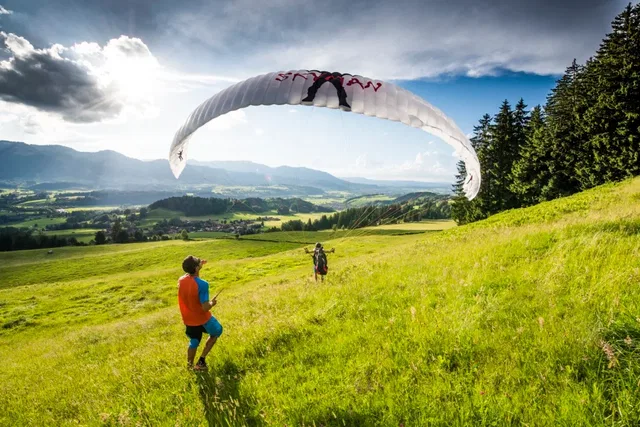 Paragliding-Szene auf grünem Hang; zwei Personen beobachten, Paraglider in der Luft über der Wiese.