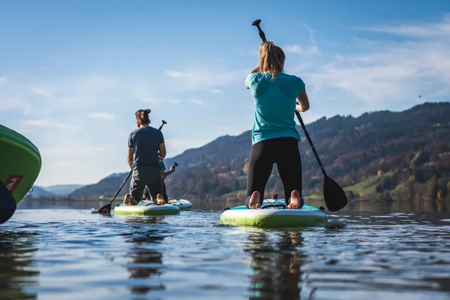 Zwei Personen stehen auf Stand-Up-Paddleboards auf einem ruhigen See, Berge im Hintergrund.