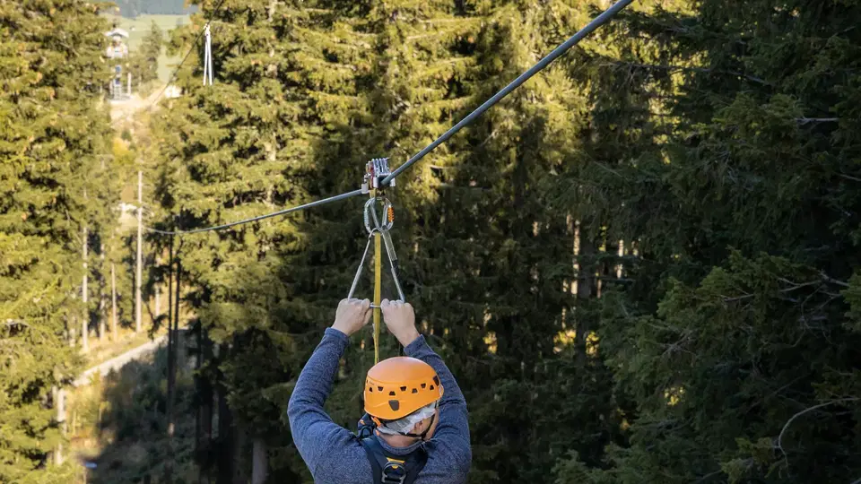 Person beim Ziplining mit Helm und Klettergurt durch Wald, Berglandschaft im Hintergrund.