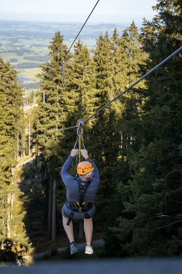 Person beim Ziplining mit Helm und Klettergurt durch Wald, Berglandschaft im Hintergrund.
