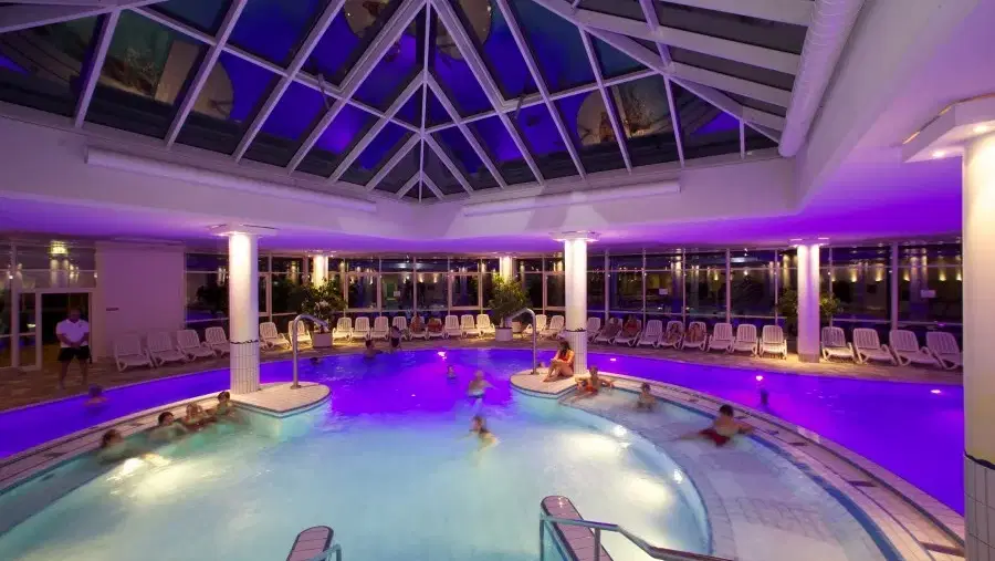 Beheiztes Innenbad mit kreisförmigem Pool, Glasdach, umlaufenden Sitzgelegenheiten und lila Beleucht