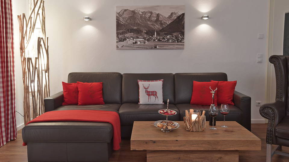 Wohnzimmer der Ferienwohnung Ifen mit dunklem Ecksofa, roten Kissen und Holztisch.