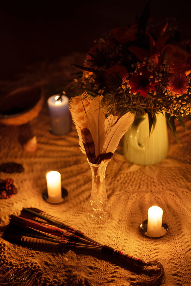 Auf einem sandfarbenen Tuch stehen mehrere brennende Kerzen, ein Blumenarrangement und eine Vase mit Federn. Daneben liegt ein dekorativer Fächer mit bunten Federn.