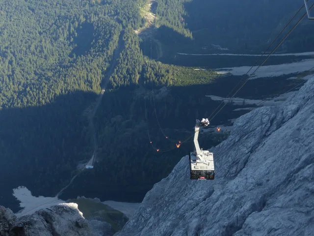 Seilbahn-Gondel schwebt über felsigem Berghang mit Wald im Hintergrund.