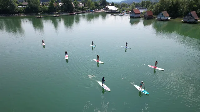 Mehrere Stand-Up-Paddler auf einem ruhigen See, Ufer mit Bäumen und Holzhausgebäuden.