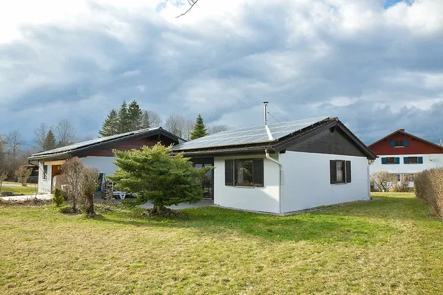 Einfamilienhaus mit weißer Fassade, dunklem Dach und Solarpanelen, von Garten umgeben.