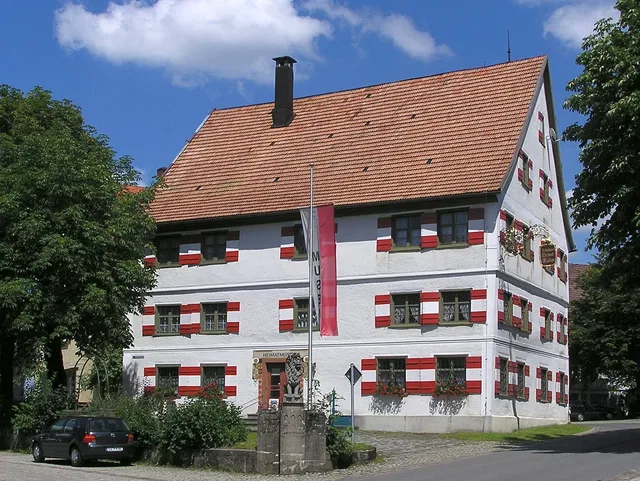 Weißes Haus mit roten Fensterläden und rotem Ziegeldach am Dorfplatz