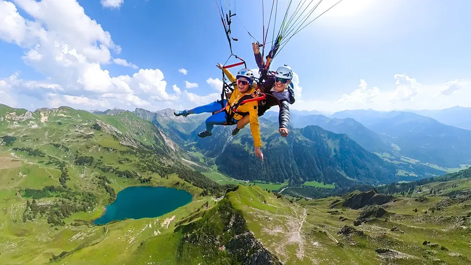 Gast und Pilot beim Tandemflug über einem Bergsee und grünen Hängen.