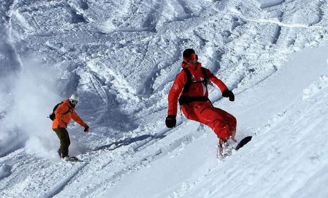 Zwei Wintersportler snowboarden eine Schneepiste hinab, links in Orange, rechts in Rot.