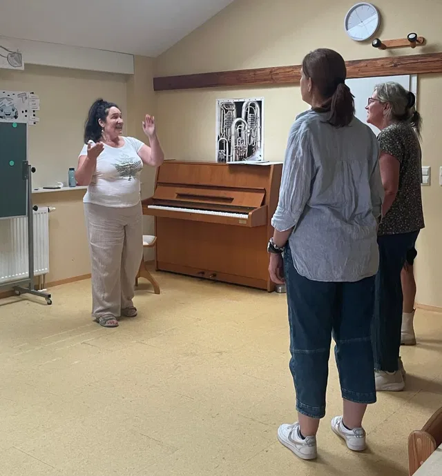 Eine Frau spricht vor drei Zuhörerinnen in einem Musikraum mit Klavier im Hintergrund.