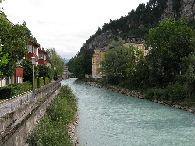 Fluss mit türkisfarbenem Wasser fließt durch eine Stadt, links Uferweg mit Häusern, rechts Ufer mitB