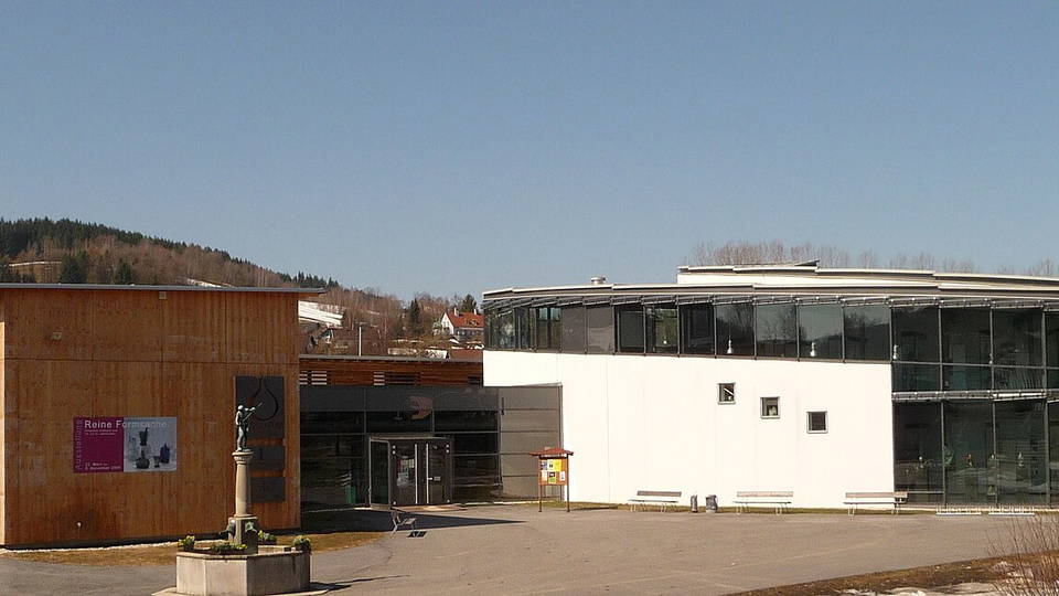 Außenansicht des Glasmuseums Frauenau mit verglaster Fassade und Vorplatz.