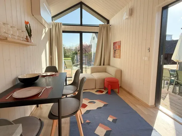 Wohnbereich eines Ferienhauses mit Esstisch, Sitzecke und großen Giebelfenstern sowie Terrassentür.