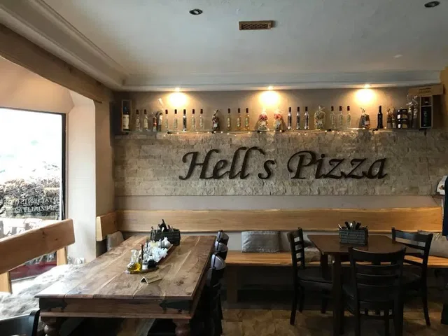Innenraum von Hell's Pizza: Holztische und Bänke, Steinwand mit Flaschenregal hinter der Bar.