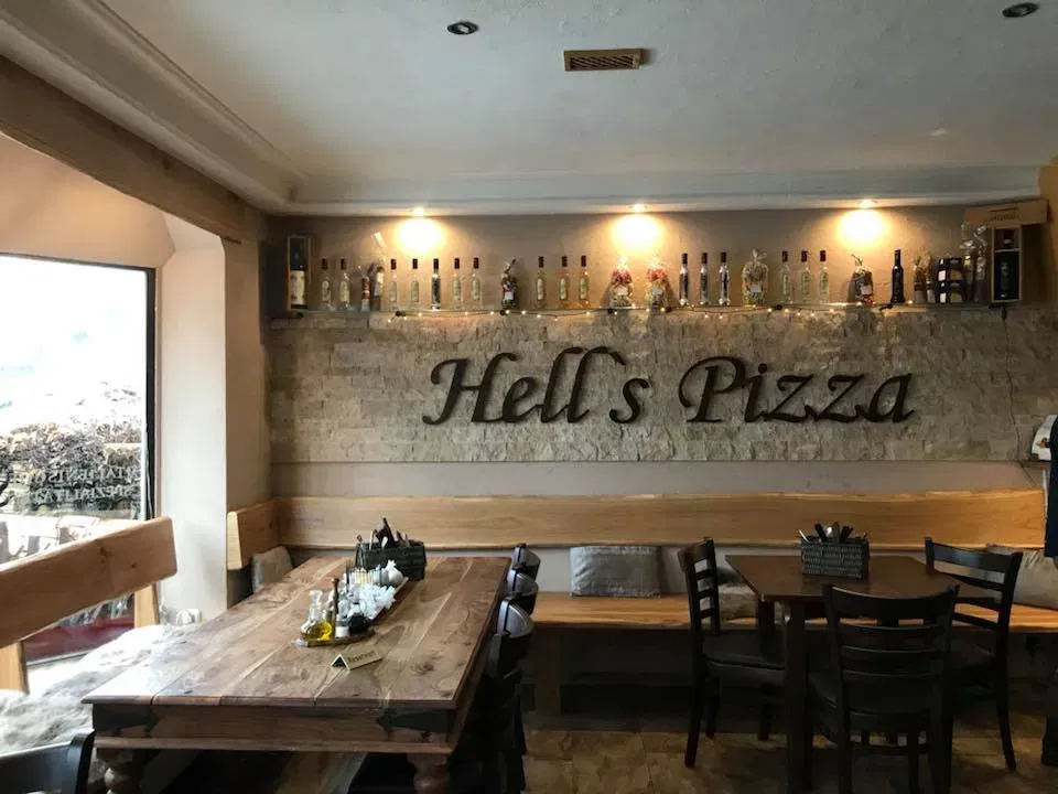 Innenraum von Hell's Pizza: Holztische und Bänke, Steinwand mit Flaschenregal hinter der Bar.