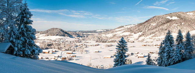 Winterliches Panorama von Hittisau mit Dorf, verschneiten Hügeln und Tannen.