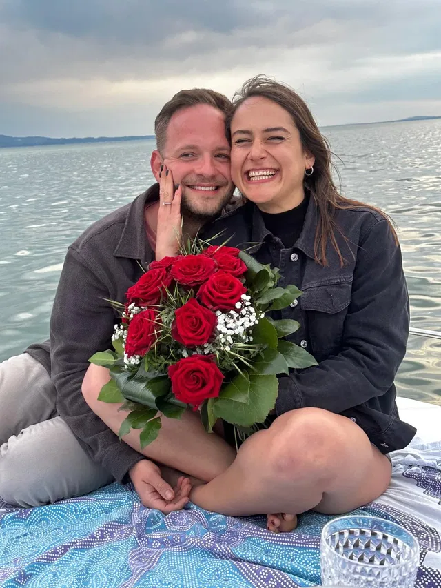 Paar mit Rosenstrauß sitzt gemütlich auf Boot am See, lächelt glücklich.