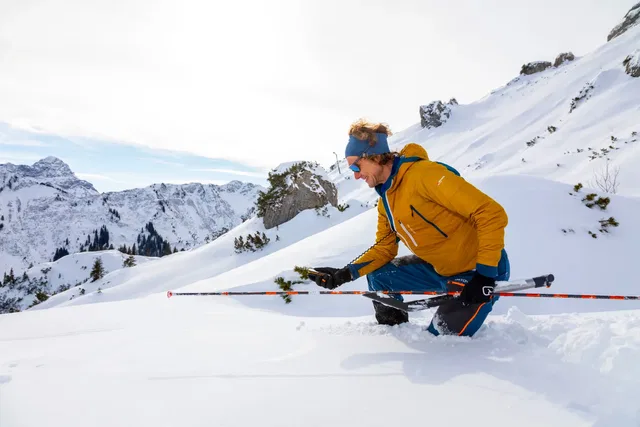 Person in gelber Winterjacke kniet im Schnee und hält Skistöcke vor bergiger Winterlandschaft.