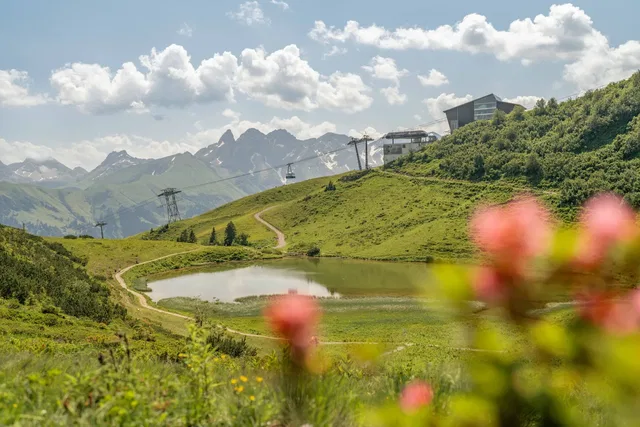 Landschaft mit grünem Hügel, kleinem See, Wanderweg, Gondelbahn und Gebäuden auf der Anhöhe.