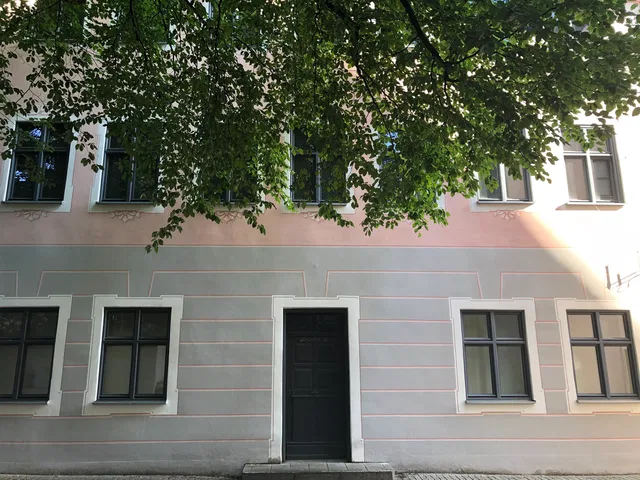 Fassade eines Gebäudes mit Fensterfront und dunkler Eingangstür, überragt von Baumkronen.