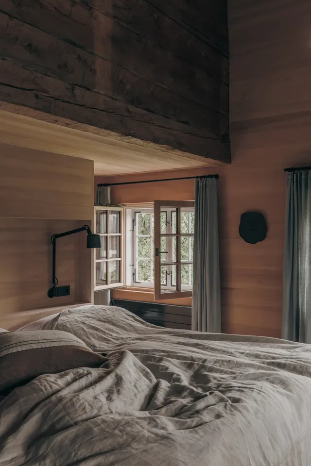 Gemütliches Schlafzimmer mit offenem Fenster, Holzbalken und Bett mit grauer Bettwäsche.