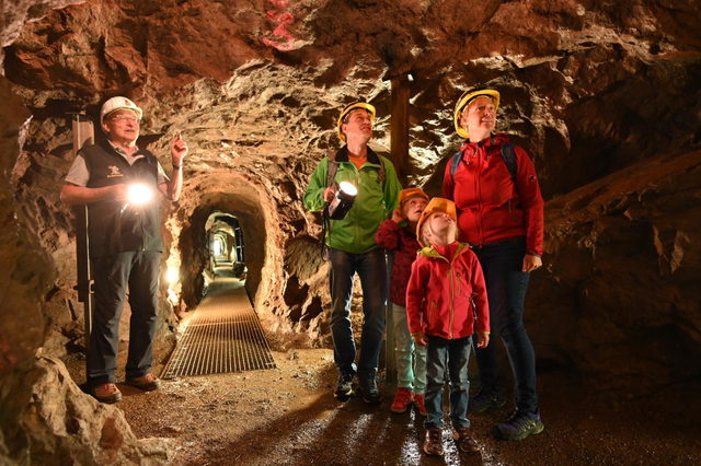 Familie mit Helm und Taschenlampe geht mit einem Grubenführer durch einen beleuchteten Stollen in den Erzgruben Burgberg.