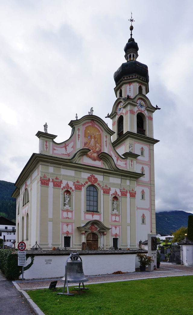 Barocke Kirche mit rosafarbener Fassade, Uhrturm und Glocke vor bewaldeten Bergen