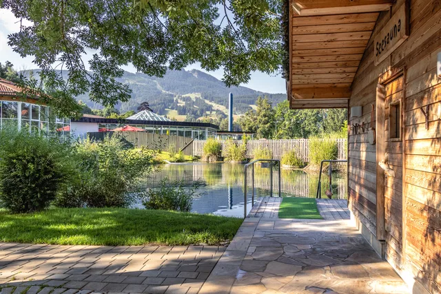 Blick auf Teich, Holzhaus und Gartenbereich mit Treppe und Mauern, Berge im Hintergrund.