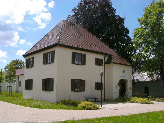 Weißes zweistöckiges Gebäude mit braunem Dach, dunklen Fensterläden und umliegendem Grün