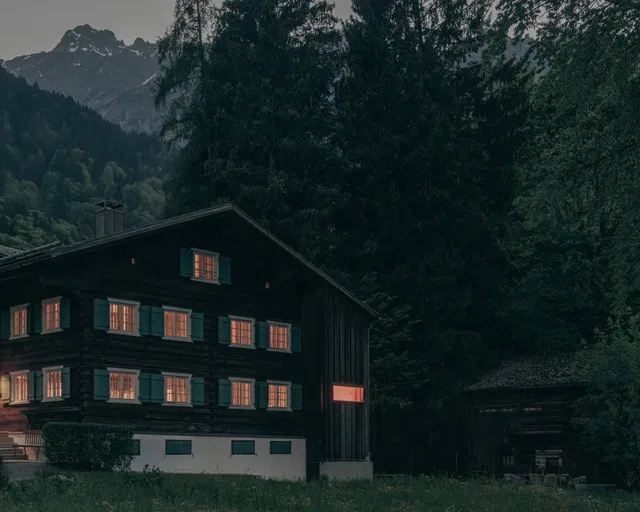 Dunkles Holzhaus im Alpenwald mit beleuchteten Fenstern bei Abenddämmerung.