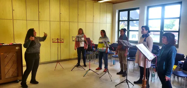 Lehrerin dirigiert Gesangsgruppe während eines Workshops mit Notenständern in einem Probenraum.