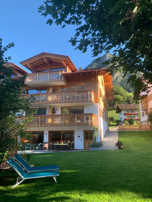 Mehrstöckiges Holz-Chalet mit Balkonen, Grünfläche und Liegestühlen vor einem Bergdorf