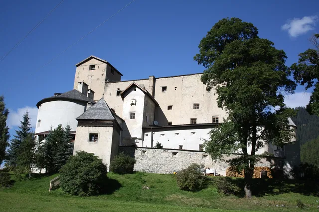 Weiße Burganlage mit Türmen auf Grünland, von Bäumen umgeben, blauer Himmel
