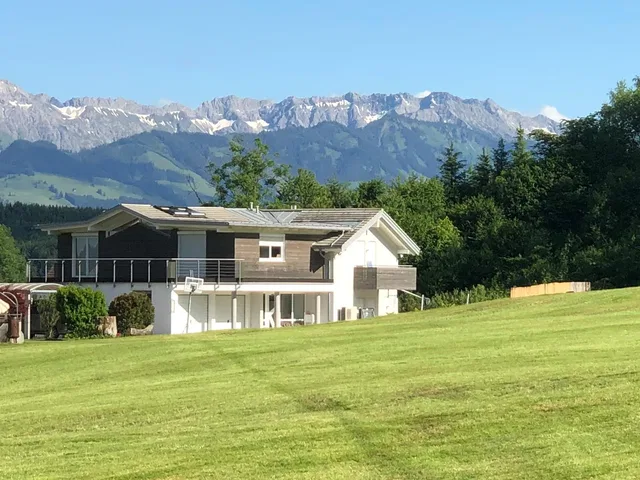 Modernes Haus auf grüner Wiese mit Alpen im Hintergrund.
