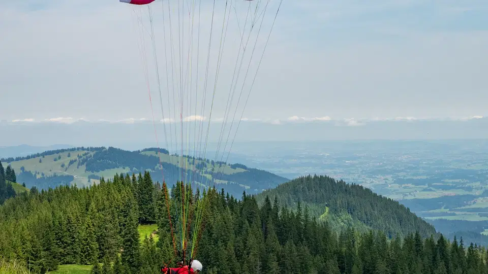 Paraglider mit rotem und weißem Schirm startet am Hang über grünem Wald.