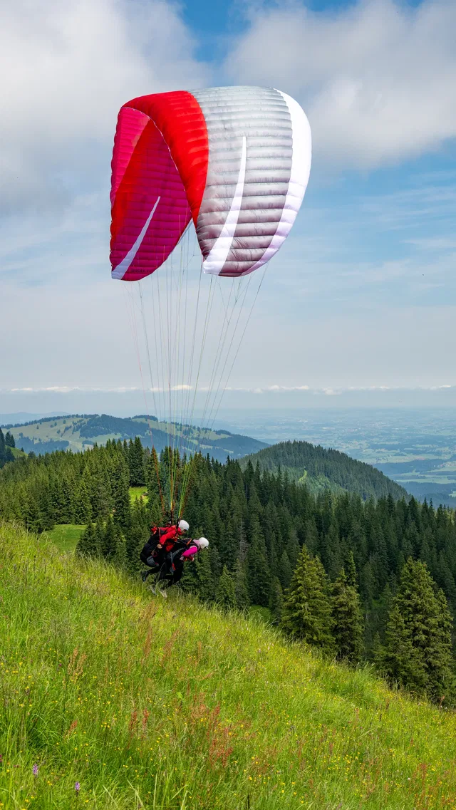 Paraglider mit rotem und weißem Schirm startet am Hang über grünem Wald.