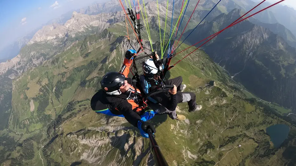 Pilot und Passagier im Tandem-Gleitschirm vor einer alpinen Berglandschaft.