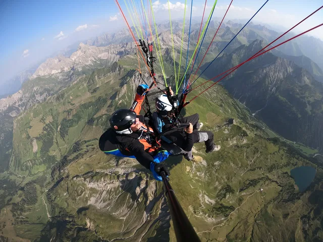 Pilot und Passagier im Tandem-Gleitschirm vor einer alpinen Berglandschaft.