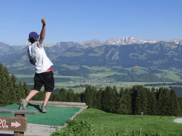 Person auf Holzplattform wirft Frisbee vor Alpenlandschaft.