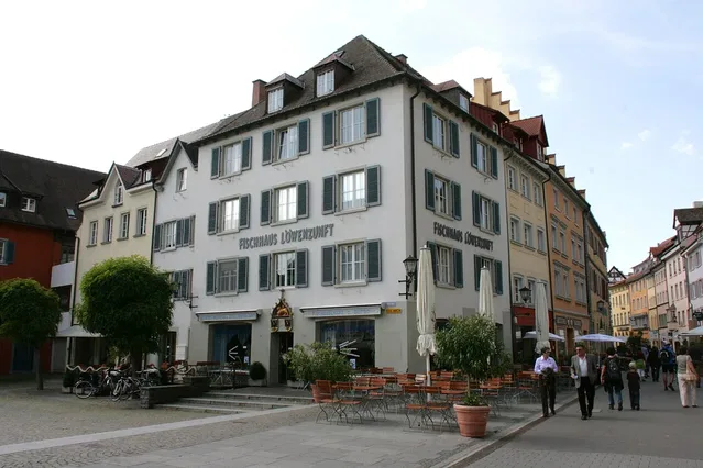 Weißes mehrstöckiges Gebäude mit grünen Fensterläden und Straßencafé vor einem historischen Platz