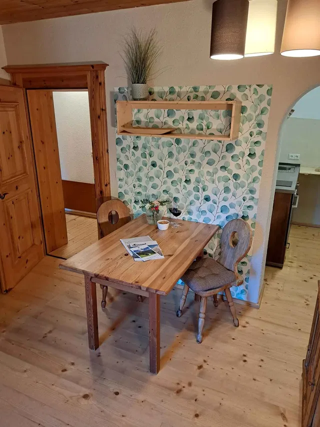 Rustikaler Holztisch mit zwei Stühlen vor Blatt-Wandtapete in einer Ferienwohnung.