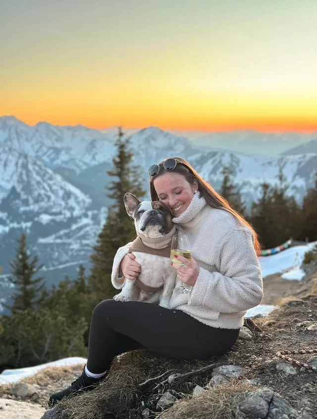 Frau knuddelt Bulldog vor Berglandschaft beim Sonnenuntergang, hält Weinglas.