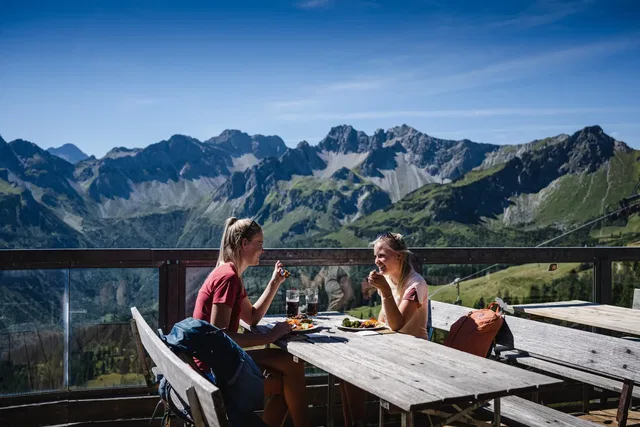 Zwei Frauen essen auf einer Bergterrasse mit Blick auf alpine Gipfel.