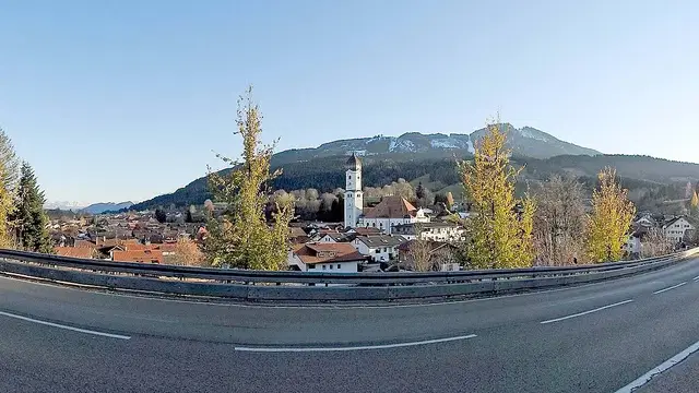 Blick über Nesselwang mit Kirchturm, herbstlichen Bäumen, Alpspitz und Edelsberg; im Vordergrund die Kemptener Straße. Ortsmotiv.