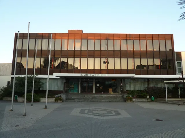 Modernes zweistöckiges Gebäude mit Glasfront, Eingangstreppe und Flaggenpfosten davor.