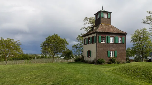 Historischer Bruchstein-Turm mit grünen Fensterläden vor Weingarten und Wolkenhimmel.