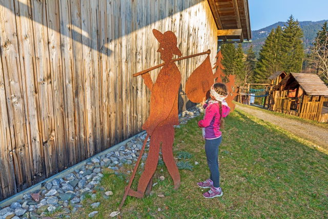 Mädchen steht im Museumsdorf Erzgruben Burgberg vor einer Knappenfigur aus Metall an einer Holzhütte.